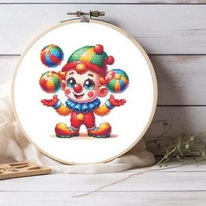 May include: A cross-stitch embroidery piece in a round wooden hoop featuring a colourful clown juggling three multi-coloured balls. The clown wears a green, yellow, and red hat, a blue ruffled collar, and a red outfit with yellow accents.