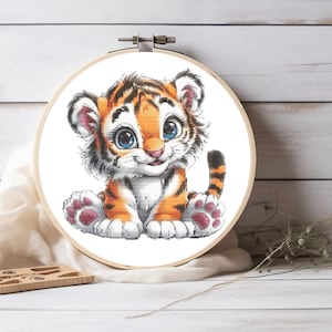 May include: A cross-stitch embroidery project featuring a cartoon tiger cub with orange fur, black stripes, and large blue eyes, secured within a round wooden embroidery hoop. The design is centered on white fabric, showcasing detailed stitching.