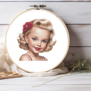 Peut inclure: Un motif de point de croix représentant une jeune fille aux cheveux blonds coiffés en style années 1950. Elle porte une robe rouge et blanche et a une fleur rouge dans les cheveux.
