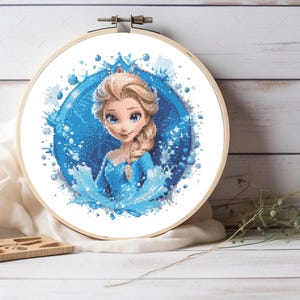 May include: A circular wooden embroidery hoop containing a cross-stitch pattern of a character with blonde hair in a braid, wearing a blue dress. The design is set against a blue circular background with white and blue ice-like crystal accents.