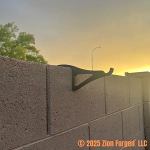 Könnte beinhalten: Ein schwarzer Metallbügel an einer grauen Betonmauer befestigt. Der Bügel hat einen gebogenen Haken und eine dreieckige Halterung. Der Hintergrund zeigt einen Sonnenuntergang, eine Straßenlaterne und Bäume. © 2025 Zion Forged LLC.