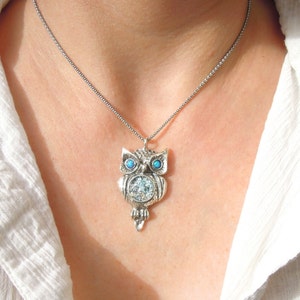Könnte beinhalten: Eine silberne Eulen-Anhänger-Kette mit blauen Opal-Augen. Die Eule ist mit einer strukturierten Oberfläche und einem runden Opal-Stein in der Mitte ihrer Brust verziert.