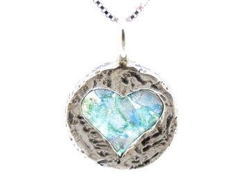 May include: A silver pendant necklace with a round, textured silver frame and a heart-shaped piece of blue and green Roman glass inside.
