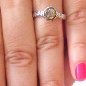 K&ouml;nnte beinhalten: Ein silberner Ring mit einem geh&auml;mmerten Band und einem runden, gefassten, braunen und goldenen Edelstein.