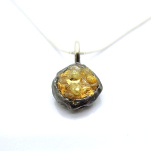 May include: A pendant necklace with an unusual, uneven pendant. The pendant has a dark, textured edge, holding a collection of yellow-gold gemstones and gold leaf. The necklace chain is a fine silver colour.