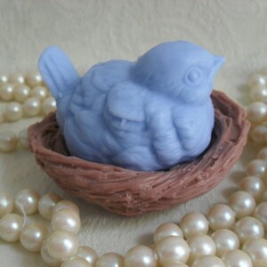 May include: A light blue bird soap sculpture sitting in a brown nest. The bird is detailed with feathers and a beak. The nest is made of woven material.