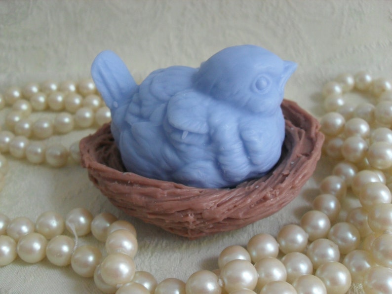 May include: A light blue bird soap sculpture sitting in a brown nest. The bird is detailed with feathers and a beak. The nest is made of woven material.