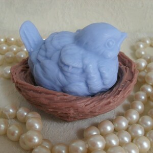 May include: A light blue bird-shaped soap bar is nestled in a brown, woven nest. The nest is surrounded by a string of white pearls.