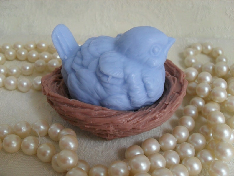 May include: A light blue bird-shaped soap bar is nestled in a brown, woven nest. The nest is surrounded by a string of white pearls.