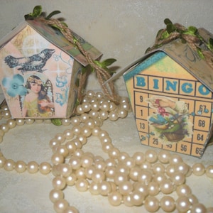 May include: Two small papercraft houses decorated with vintage paper and embellishments. One house features a blue bird and a girl with wings, the other has a bingo card with a bird and a basket of flowers. Both houses are adorned with twine and green leaves.