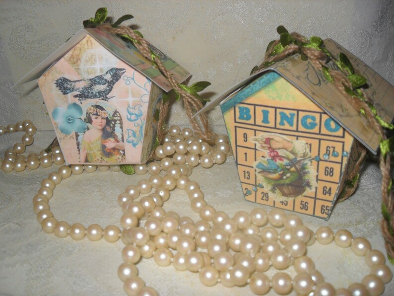 May include: Two small papercraft houses decorated with vintage paper and embellishments. One house features a blue bird and a girl with wings, the other has a bingo card with a bird and a basket of flowers. Both houses are adorned with twine and green leaves.