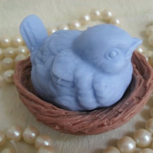 May include: A light blue bird figurine nestled in a brown woven nest. The bird has a detailed face and is positioned as if it is sleeping.