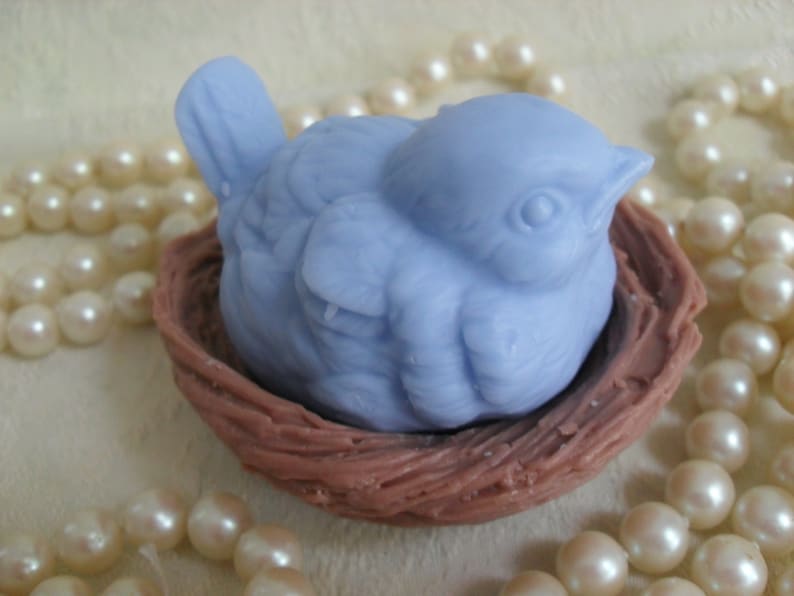 May include: A light blue bird figurine nestled in a brown woven nest. The bird has a detailed face and is positioned as if it is sleeping.