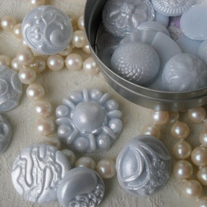 May include: A collection of pearl beads and silver-toned buttons with intricate designs. The buttons are arranged in a circular tin container and on a white surface.