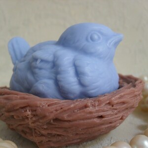 May include: A light blue bird-shaped soap bar is nestled in a brown, woven nest. The nest is made of a textured material that resembles twigs.