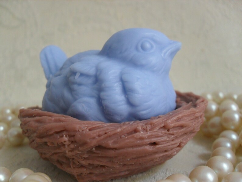 May include: A light blue bird-shaped soap bar is nestled in a brown, woven nest. The nest is made of a textured material that resembles twigs.