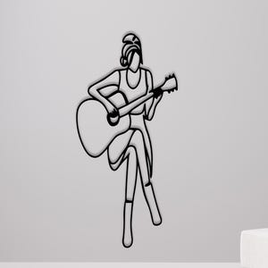 May include: Black metal wall art of a person playing an acoustic guitar. The artwork is a minimalist line drawing, mounted on a light grey wall. The room includes a chair, footstool, and side table, creating a modern aesthetic.