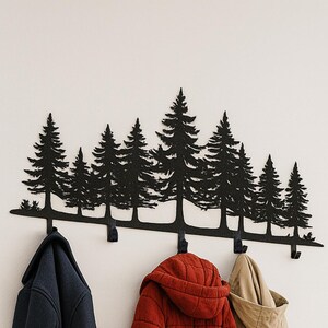 Puede incluir: Un perchero de pared de metal negro con la silueta de pinos. Tres abrigos en azul marino, rojo y beige cuelgan de los ganchos. El perchero está montado en una pared blanca cerca de una puerta blanca.