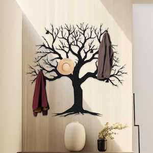 May include: Black metal tree wall hook with branches for hanging coats, hats, and other items. The hook is mounted on a beige wall with a wooden shelf and a vase with a plant.