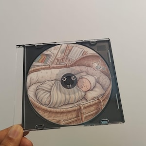 Peut inclure: Un boîtier de CD contenant un CD avec une illustration d'un bébé endormi dans un berceau en osier. Le bébé est enveloppé dans une couverture à rayures et porte un bonnet en tricot. Un petit oiseau est perché sur le côté.
