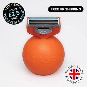 May include: An orange spherical razor with a multi-blade cartridge head. The handle is a round ball shape with the embossed text RAZABALL. The razor head features a grey plastic housing with a blue lubricating strip and a small smiley face icon.