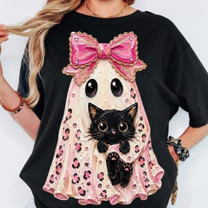May include: A black t-shirt featuring a graphic of a ghost wearing a large pink bow and holding a black kitten. The ghost has a light pink leopard print pattern and is decorated with small rhinestones throughout the design.