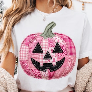 May include: A white t-shirt featuring a large, central graphic of a pumpkin made from a patchwork of various pink fabrics. The pumpkin has a green stem and black fabric eyes, nose, and a jagged mouth. The patterns include gingham and ribbons.
