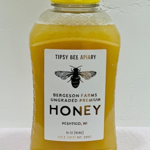 May include: A clear plastic bottle of honey with a yellow lid. The label reads "Tipsy Bee Apiary", "Bergeson Farms", "Ungraded Premium Honey", "Peshtigo, WI", "16 oz (454g)", and "426 S. Emery Ave. 54157". A black and white illustration of a bee is on the label.