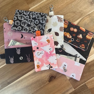 May include: Six small fabric pouches with different Halloween-themed prints. The pouches are pink, black, white, and orange with various designs including pumpkins, ghosts, and leaves. Each pouch has a metal ring and a zipper.