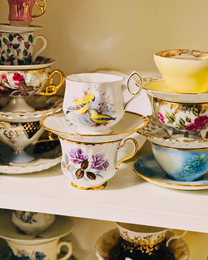 A collection of teacups and saucers with floral and bird designs. The teacups are white with gold trim and feature various floral and bird patterns. The saucers are also white with gold trim and match the teacups.