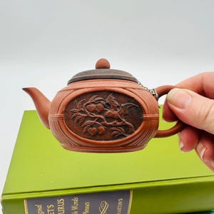 May include: A small, reddish-brown teapot with a carved design of a bird and fruit. The lid is dark brown, and a small chain connects the lid to the handle. The teapot is resting on a green book.