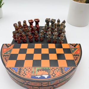 May include: A chess set featuring a semi-circular board with a black and orange checkered pattern and decorative geometric borders. The set includes two distinct groups of hand-painted, detailed figurines in red and grey tones arranged on the board.
