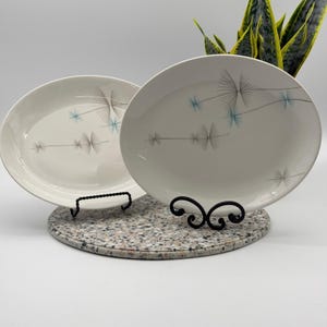 May include: Two oval-shaped white ceramic plates featuring a delicate pattern of thin, dark gray lines forming starburst or dandelion-like shapes, accented with small, light blue linear details scattered across the smooth, glossy surface.