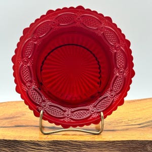 May include: A vibrant red glass dish with a scalloped edge and intricate embossed detailing. The dish features a central, textured design and is displayed on a gold-toned metal stand. A decorative piece for serving or display.