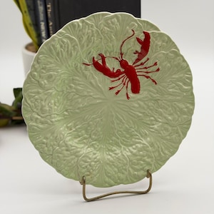 May include: A round ceramic plate with a light green, embossed cabbage leaf texture and a glossy finish. A bright red lobster is centered on the plate, featuring detailed claws and legs. The plate is supported by a thin, gold-toned metal stand.