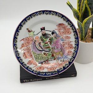 May include: A round ceramic decorative plate featuring a hand-painted scene of three figures in traditional robes standing among stylized red clouds. The plate has a wide, dark blue border with a repeating gold and black scrollwork pattern.