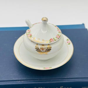 May include: A white porcelain sugar bowl with a lid and a small spoon, resting on a matching saucer. The bowl and lid are decorated with pink roses and gold accents. The sugar bowl is a vintage item.