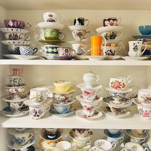 May include: A collection of vintage teacups and saucers displayed on white shelves. The teacups feature various floral patterns, gold trim, and unique designs. The arrangement includes cups of different sizes and colors, creating a charming display.