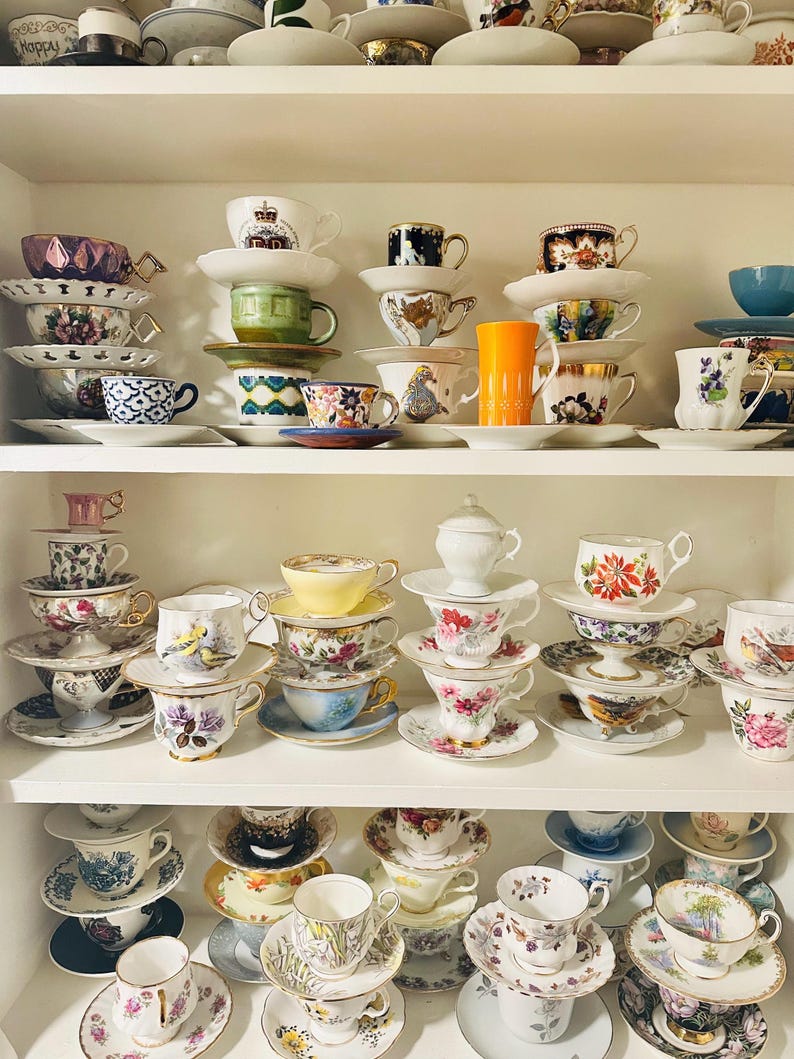 A white 3 tier shelf filled with a beautiful collection of teacups and saucers. The teacups are of various colors and designs, including floral, geometric, and plain white. Some of the teacups and saucers are stacked on top of each other. The designs vary from classic Made in England bone china to modern designs.