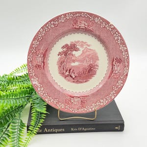 May include: A round ceramic plate featuring a cream-colored center with a detailed red landscape illustration of trees, mountains, and figures. The wide rim has a textured red border with repeating floral and architectural motifs.