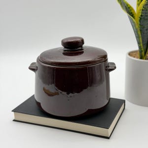 May include: A dark brown ceramic pot with a matching lid and two side handles. The pot has a smooth, glossy finish and a rounded body shape. It is positioned on top of a rectangular book with a black cover and white pages, showing a reflective surface.