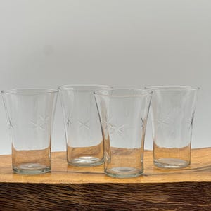 May include: Four clear drinking glasses with a starburst design etched into the glass. The glasses are cylindrical with a slight taper and sit on a wooden surface. The background is a solid, neutral color.