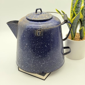 May include: A dark blue enamel coffee pot with a white speckled pattern, featuring a matching lid, a side handle with a black grip, a metal bail handle, and a small secondary handle on the side. The pot has a wide base and a tapered spout.