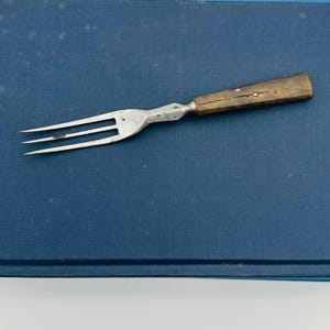May include: An antique fork with three tines and a wooden handle. The metal fork has a silver finish and shows signs of wear. The handle is made of dark wood and is attached with a metal rivet. The fork is set against a blue background.