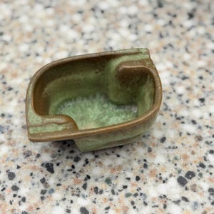 May include: A small, irregularly shaped ceramic bowl with a textured brown exterior and a smooth, mottled light green interior glaze. The vessel features a low, open-top design with thick, rounded edges and a deep, recessed basin for holding small items.