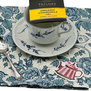 A beautiful and delicate white teacup and saucer with blue floral designs, a silver teaspoon and a tea bag  on a white and blue floral napkin. There is also a small pink teapot sticker.