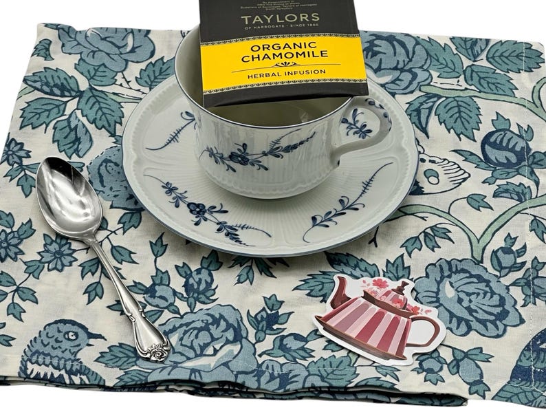 A beautiful and delicate white teacup and saucer with blue floral designs, a silver teaspoon and a tea bag  on a white and blue floral napkin. There is also a small pink teapot sticker.