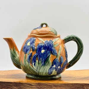 May include: A ceramic teapot with a lid, featuring a design of blue iris flowers on a tan background. The handle and spout are green, and the teapot sits on a wooden surface.