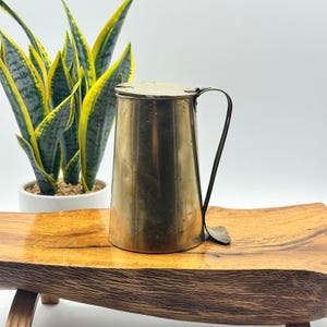 May include: A vintage brass pitcher with a lid and a curved handle. The pitcher has a tapered shape and a shiny finish. It is displayed on a wooden tray with a plant in the background. The pitcher is likely used for serving beverages.