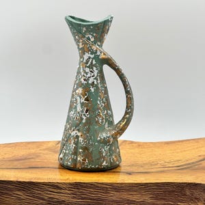 May include: A ceramic pitcher with a flared, scalloped rim and a single handle. The surface features a mottled finish in shades of sage green, white, and metallic gold, creating a textured, speckled appearance across the entire exterior body.